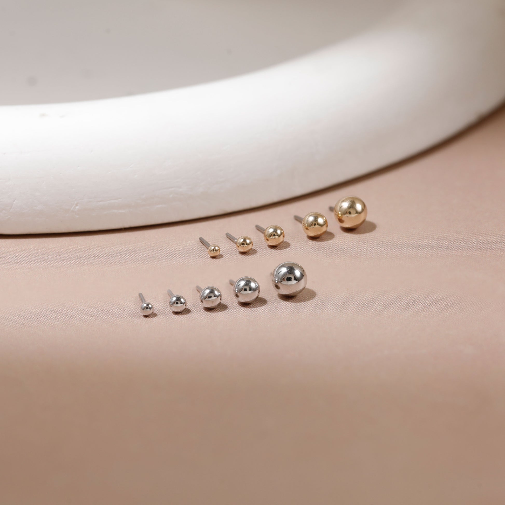 Nine small spherical stud earrings in silver and gold finishes, arranged in rows on a warm beige surface with a white curved ceramic prop in the background.