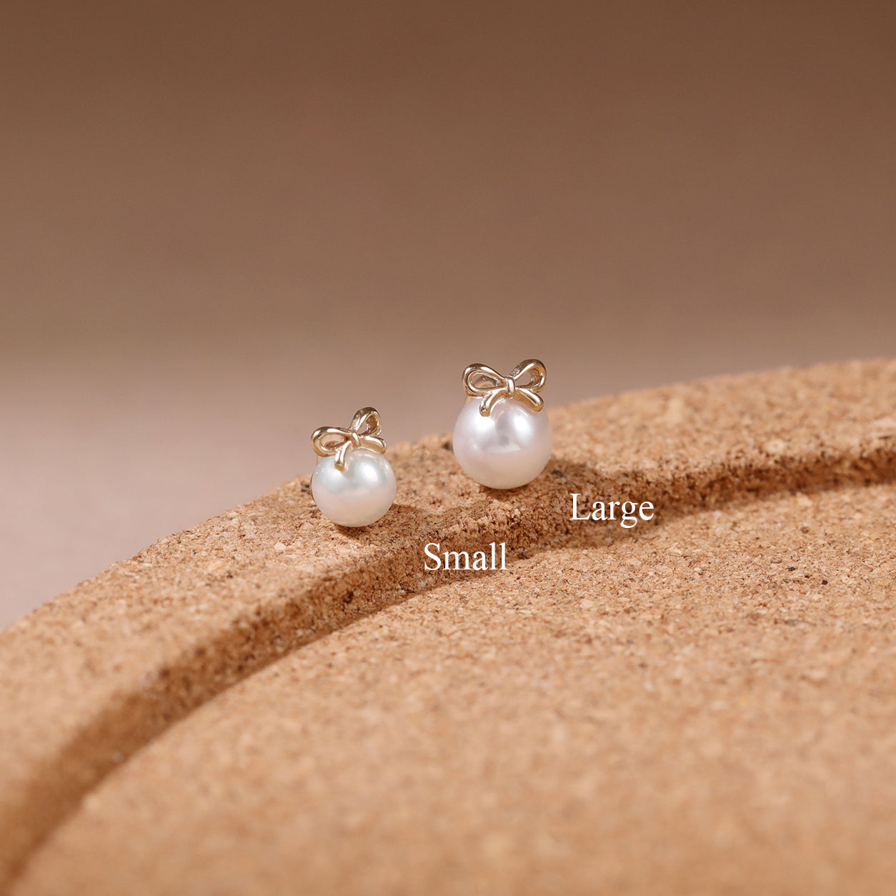 Two gold bow stud earrings with white pearl drops, labeled Small and Large, displayed on a textured brown cork-like surface against a warm beige background.