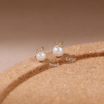 Two gold bow stud earrings with white pearl drops, labeled Small and Large, displayed on a textured brown cork-like surface against a warm beige background.