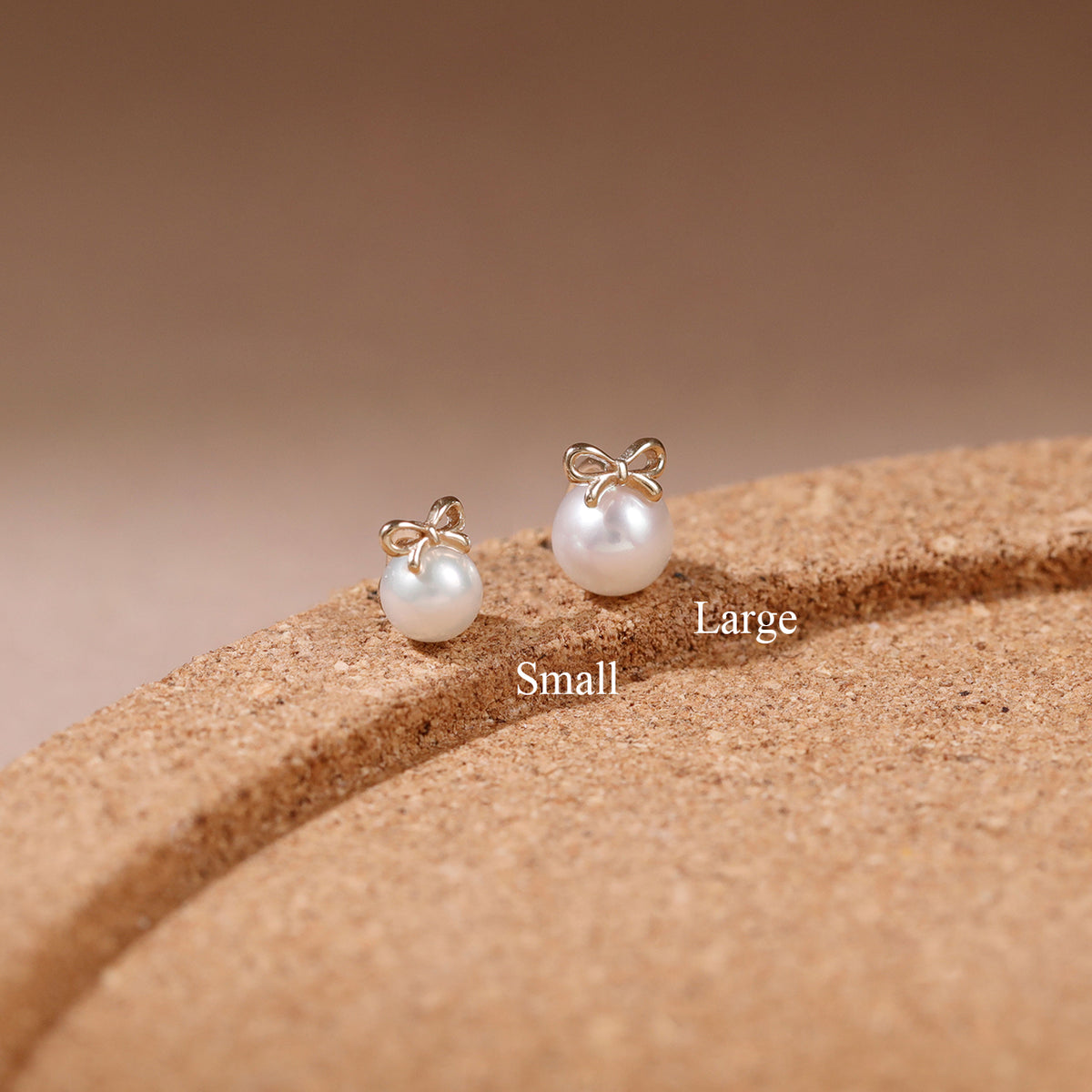 Two gold bow stud earrings with white pearl drops, labeled Small and Large, displayed on a textured brown cork-like surface against a warm beige background.