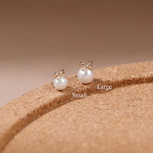 Two gold bow stud earrings with white pearl drops, labeled Small and Large, displayed on a textured brown cork-like surface against a warm beige background.