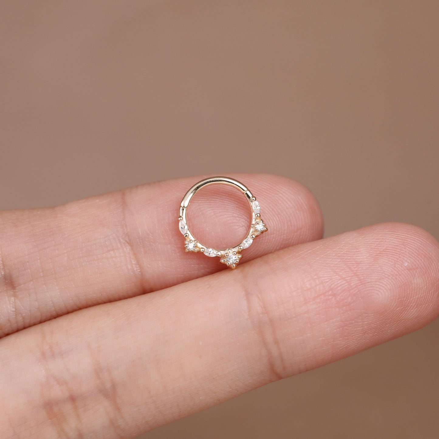 A delicate gold ring with small clear gemstones, shown resting on a person's fingers against a soft brown background.