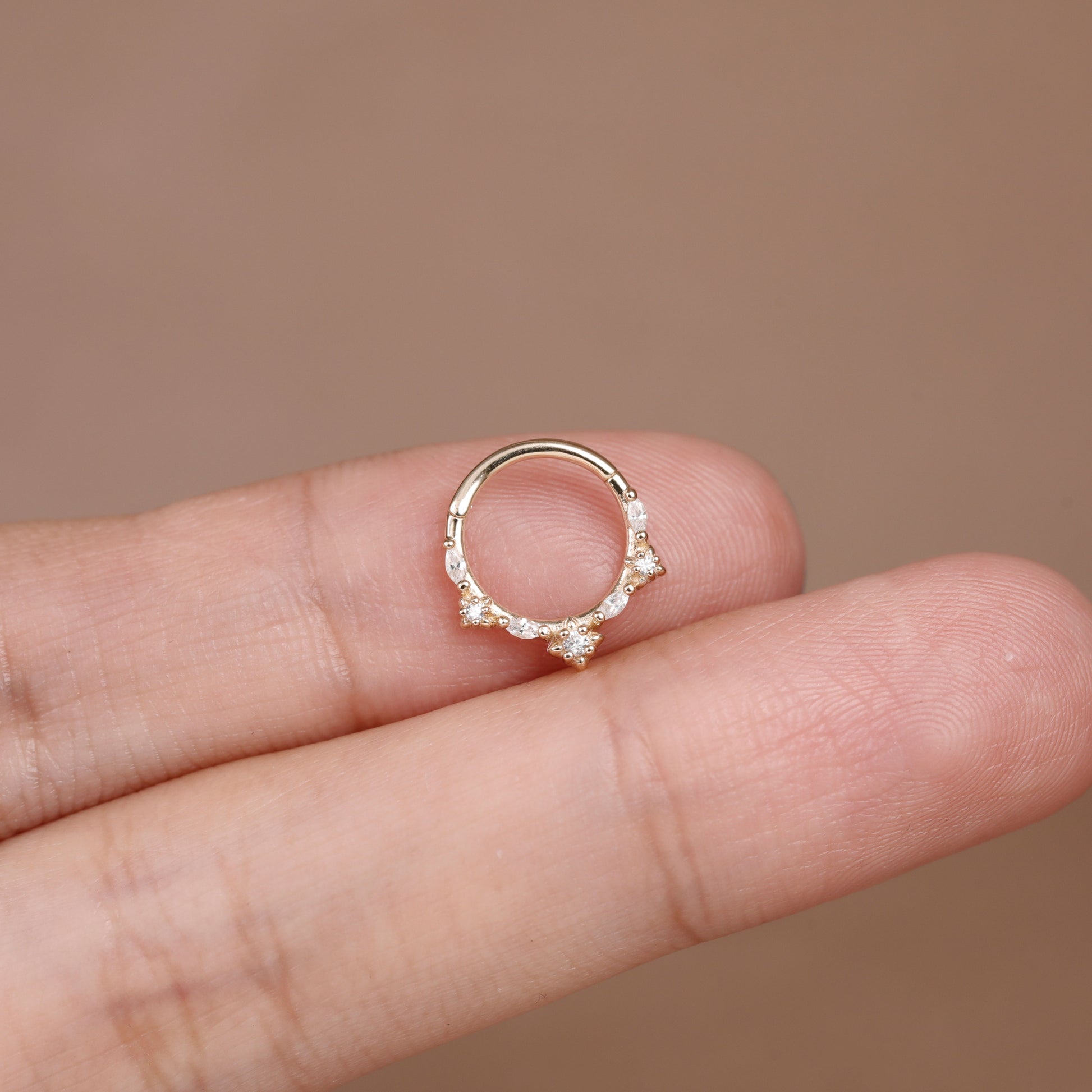 A delicate gold ring with small clear gemstones, shown resting on a person's fingers against a soft brown background.
