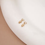 A pair of small gold hamsa hand earrings with clear pavé stones, displayed on a curved white ceramic surface.