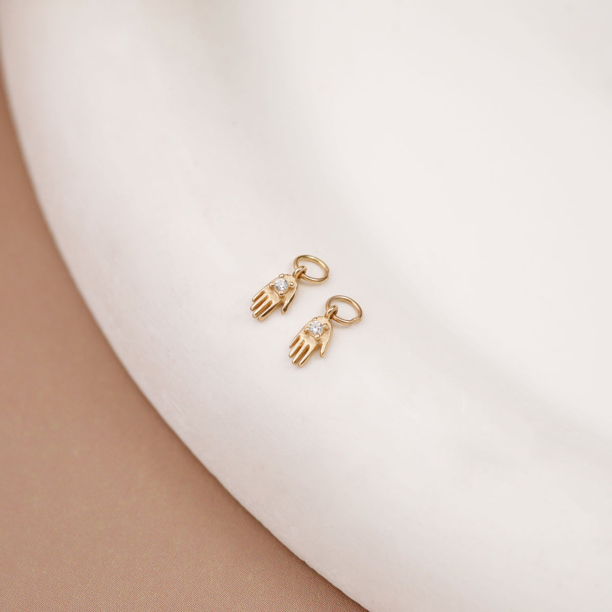 A pair of small gold hamsa hand earrings with clear pavé stones, displayed on a curved white ceramic surface.
