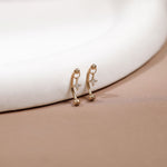 A pair of small gold huggie hoop earrings with dangling star-shaped charms, resting on a beige surface next to a white curved display stand.