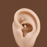 Two small gold ear piercings with clear faceted gemstones displayed on a beige ear model against a solid brown background.