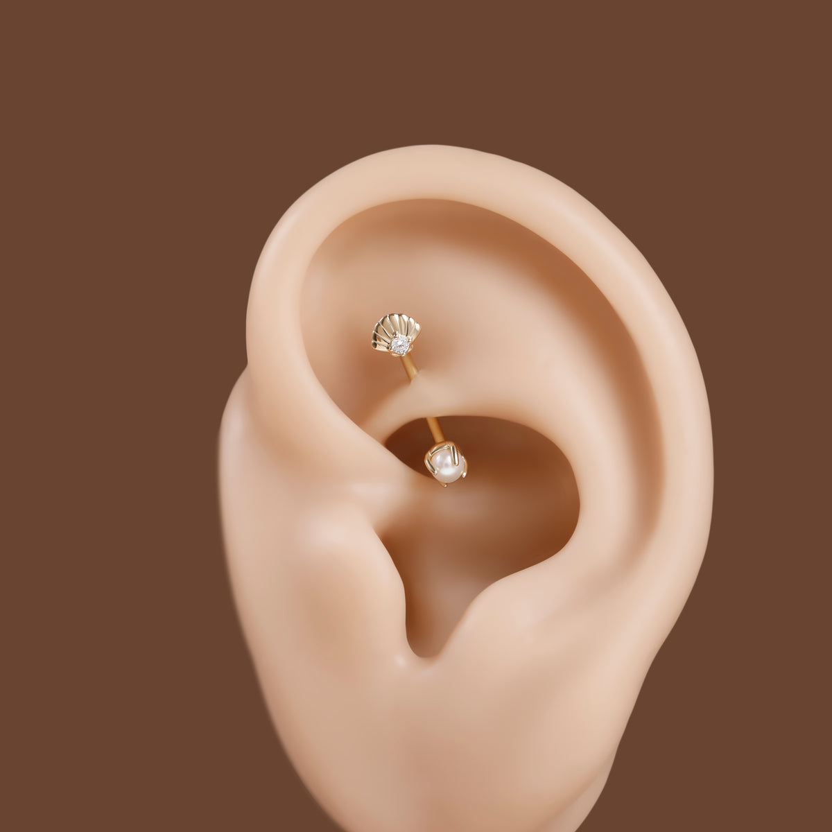 Two small gold ear piercings with clear faceted gemstones displayed on a beige ear model against a solid brown background.