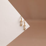 A pair of gold huggie hoop earrings with small dangling hand charms set with clear stones, displayed on a geometric white and warm brown background.