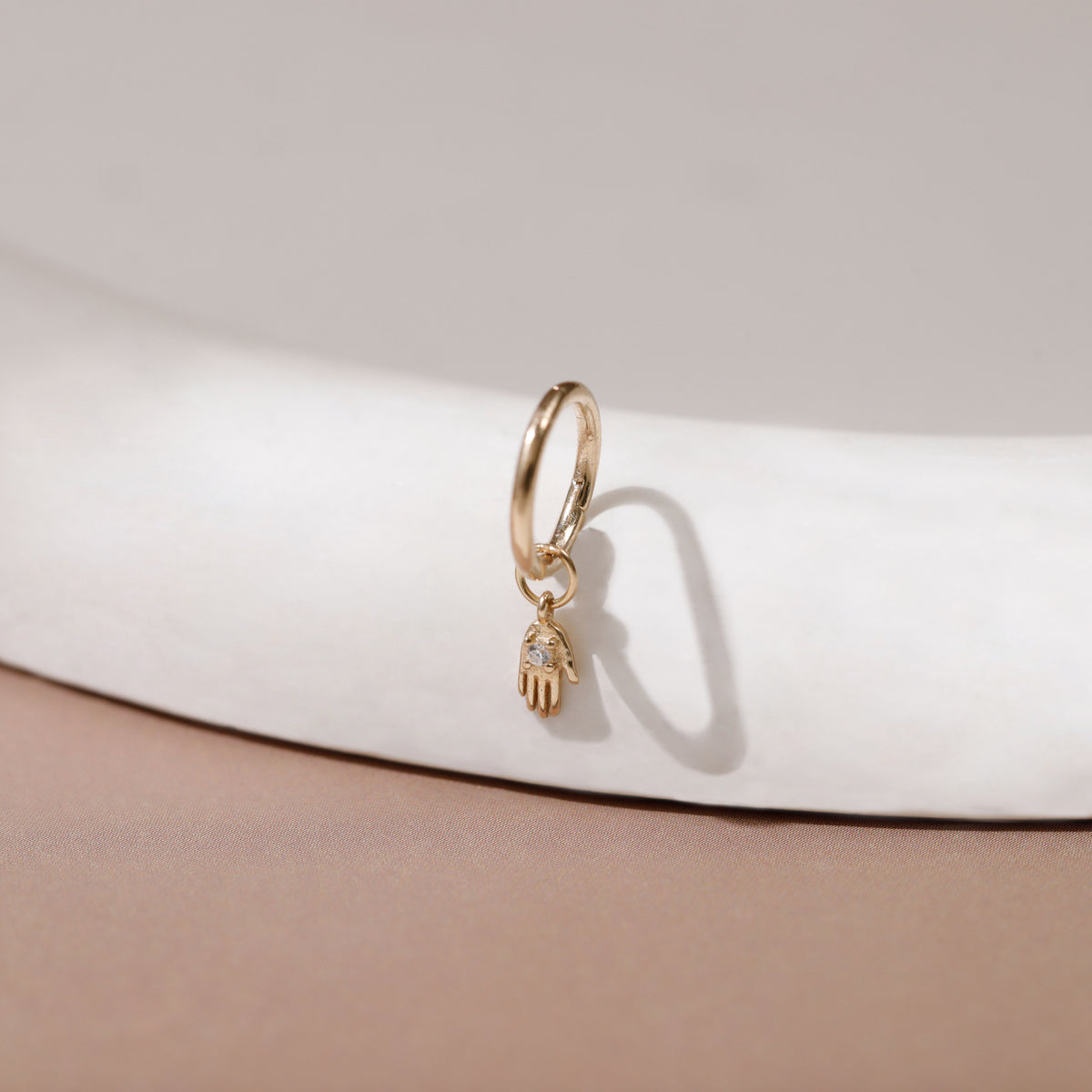 A gold huggie hoop earring with a dangling Hamsa hand charm set with small clear stones, resting on a white curved display stand against a beige background.
