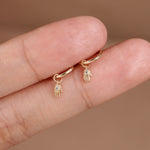 A pair of small gold huggie hoop earrings with dangling Hamsa hand charms, shown worn on a person's fingers against a soft beige background.