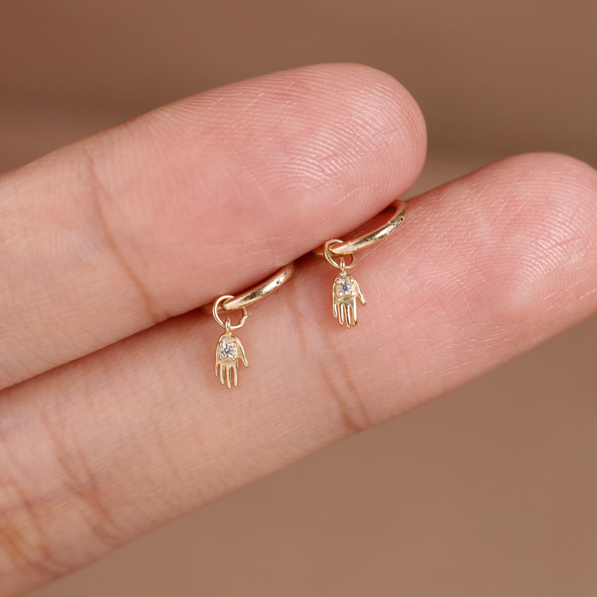 A pair of small gold huggie hoop earrings with dangling Hamsa hand charms, shown worn on a person's fingers against a soft beige background.