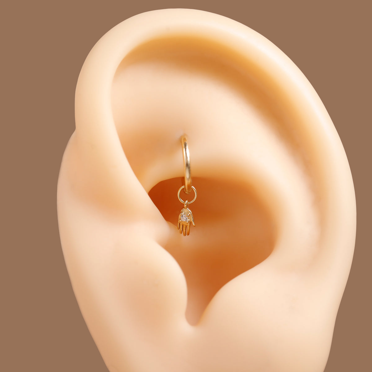 A small gold huggie hoop earring with a dangling hamsa hand charm set with a tiny clear stone, displayed on a beige ear model against a solid brown background.