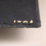Four small gold stud earrings displayed on a dark gray slate slab against a light beige background. The earrings feature simple organic shapes: a teardrop, two overlapping lotus petals, three overlapping lotus buds, and a heart.