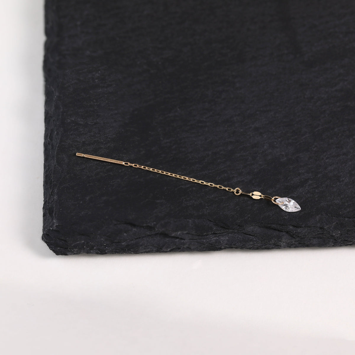 A delicate gold-toned ear cuff with a flat bar and a small clear teardrop crystal charm, displayed on a textured black slate surface.