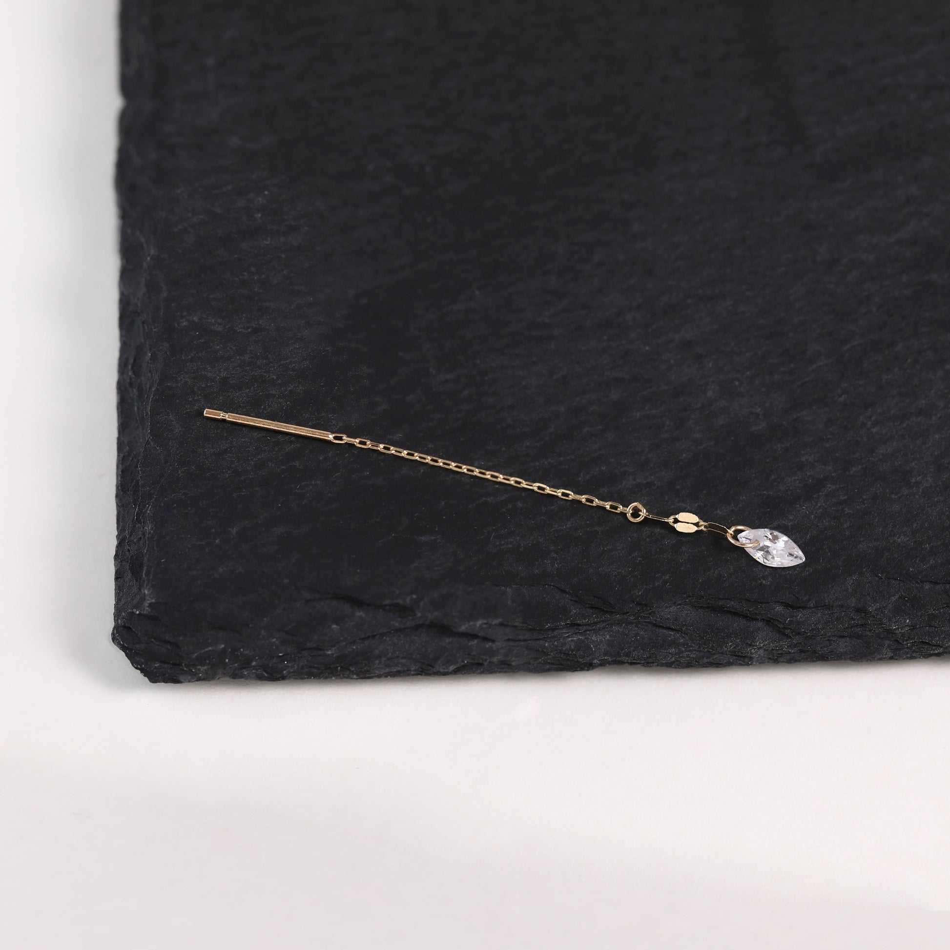A delicate gold-toned ear cuff with a flat bar and a small clear teardrop crystal charm, displayed on a textured black slate surface.