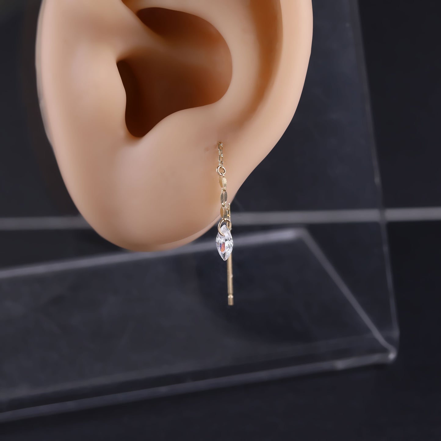 A gold ear cuff earring with a dangling clear marquise-cut crystal, displayed on a beige plastic ear mannequin against a dark background.
