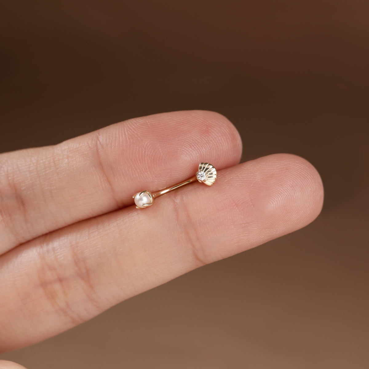 A close-up of a person's fingers holding a delicate gold ring with two small clear faceted gemstones against a warm brown background.