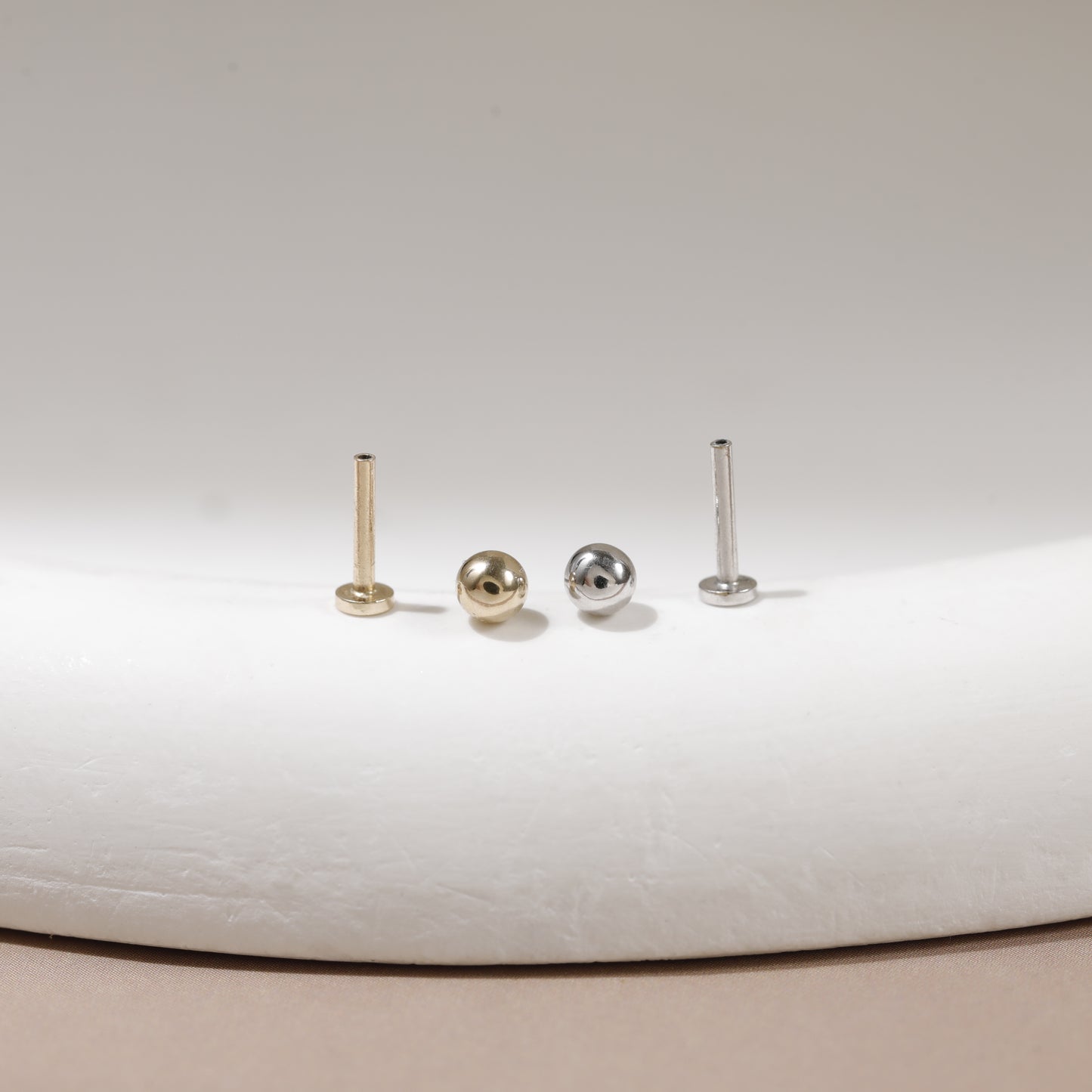 Three small stud earrings displayed on a white curved surface: two gold-toned posts and one silver-toned post with a round ball stud in the center.