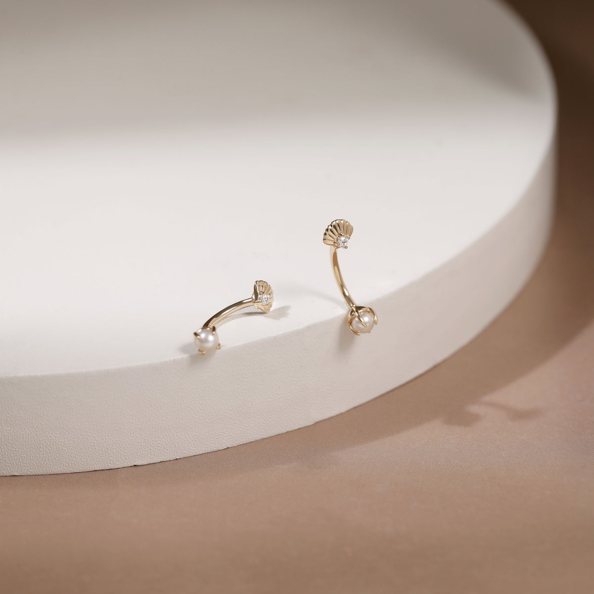 A pair of gold-toned ear cuffs with a scalloped shell design and a small pearl-like bead at the end, displayed on a white curved pedestal against a warm beige background.