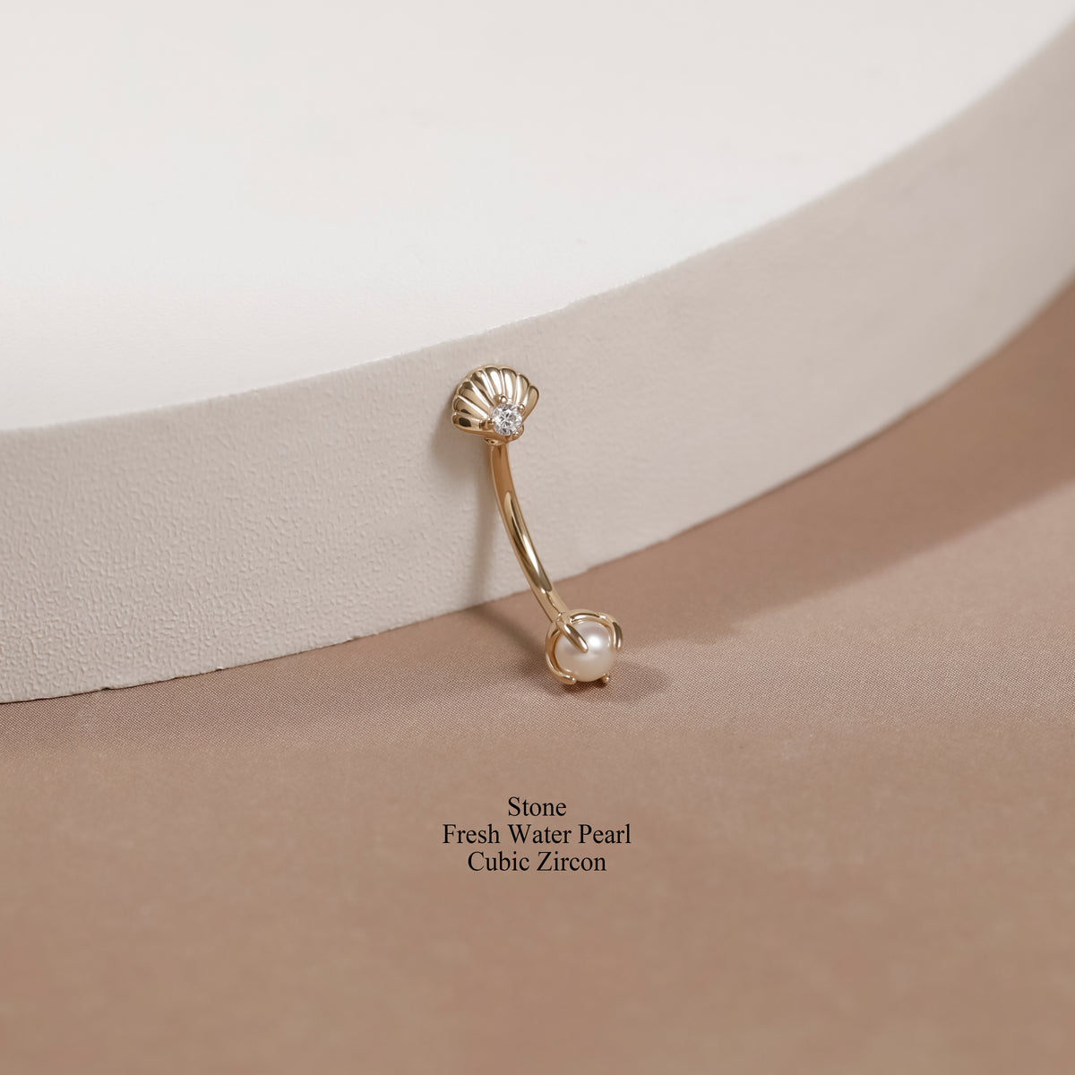 A gold-toned curved barbell navel piercing stud with a scalloped shell top set with a clear stone and a fresh water pearl dangling at the bottom, displayed on a beige surface next to a white curved pedestal.