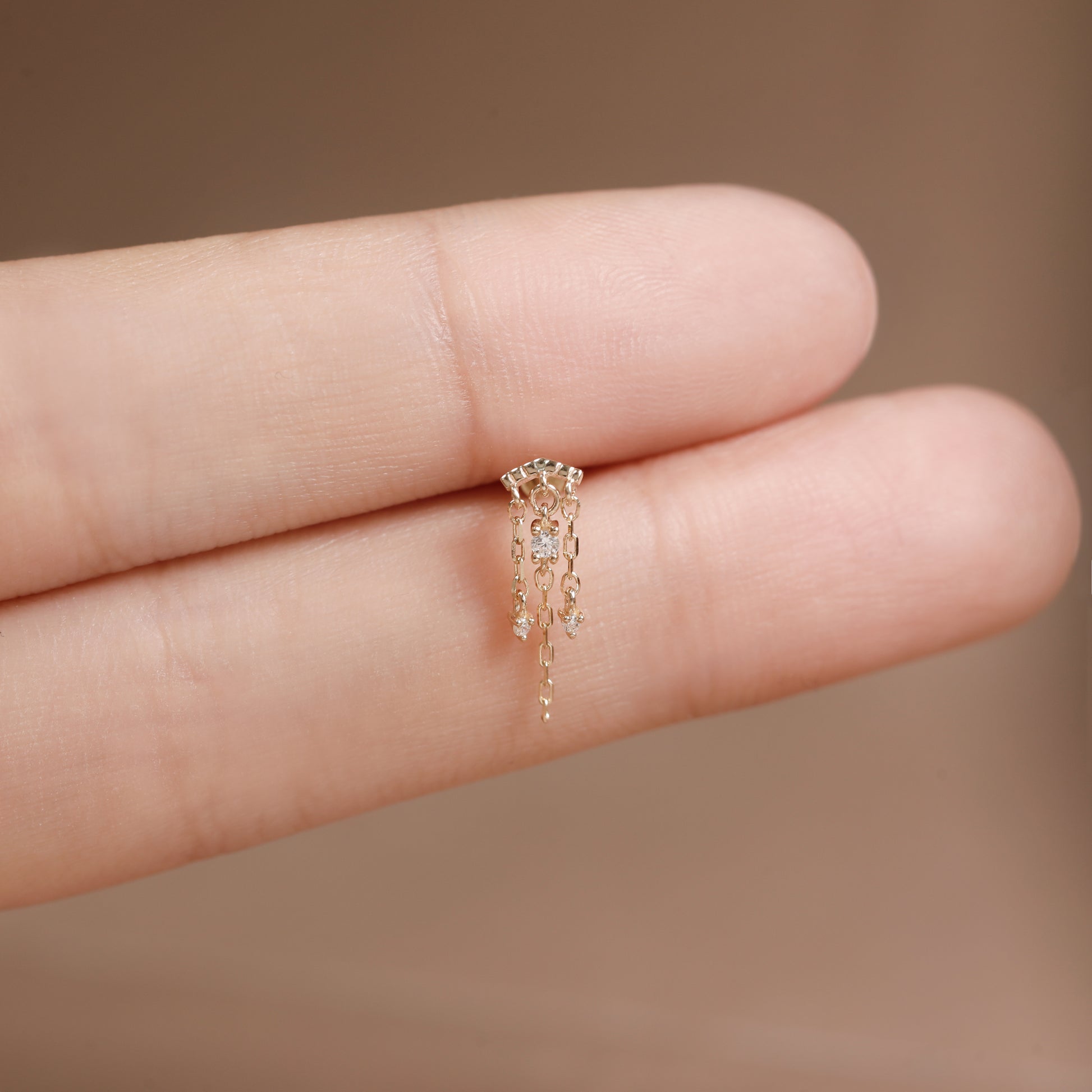 A delicate gold earring with small dangling clear stones, shown resting on a person's fingers against a soft beige background.