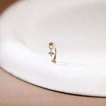 A delicate gold huggie hoop earring with a small dangling star and clear gemstone, resting on a white ceramic dish against a soft beige background.
