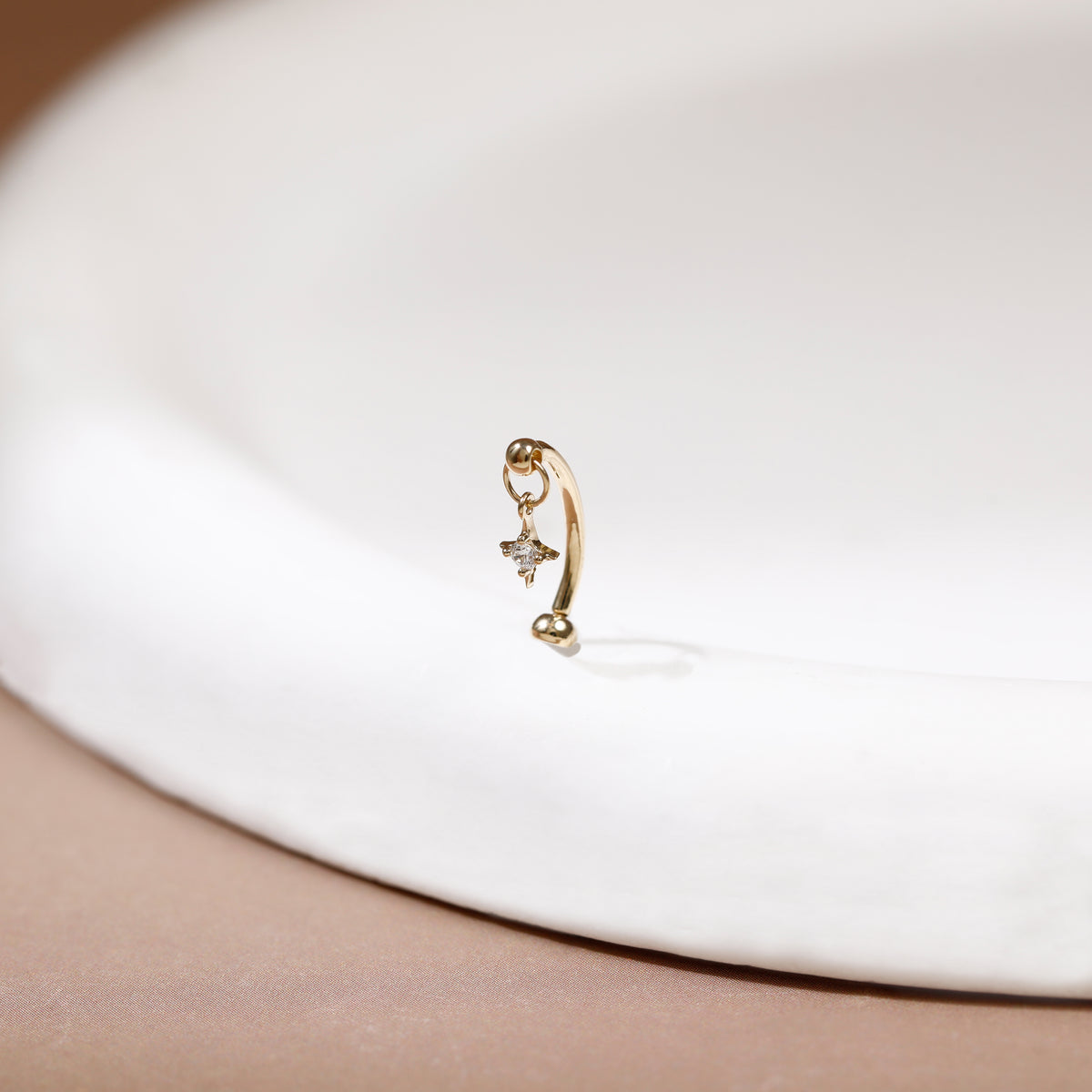 A delicate gold huggie hoop earring with a small dangling star and clear gemstone, resting on a white ceramic dish against a soft beige background.