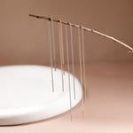 Three pairs of long dangling chain earrings in silver and gold tones, hanging from a thin branch above a white ceramic dish against a warm beige background.