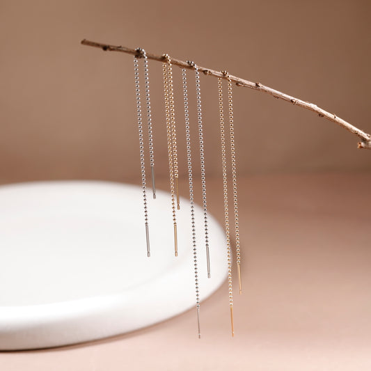 Three pairs of long dangling chain earrings in silver and gold tones, hanging from a thin branch above a white ceramic dish against a warm beige background.