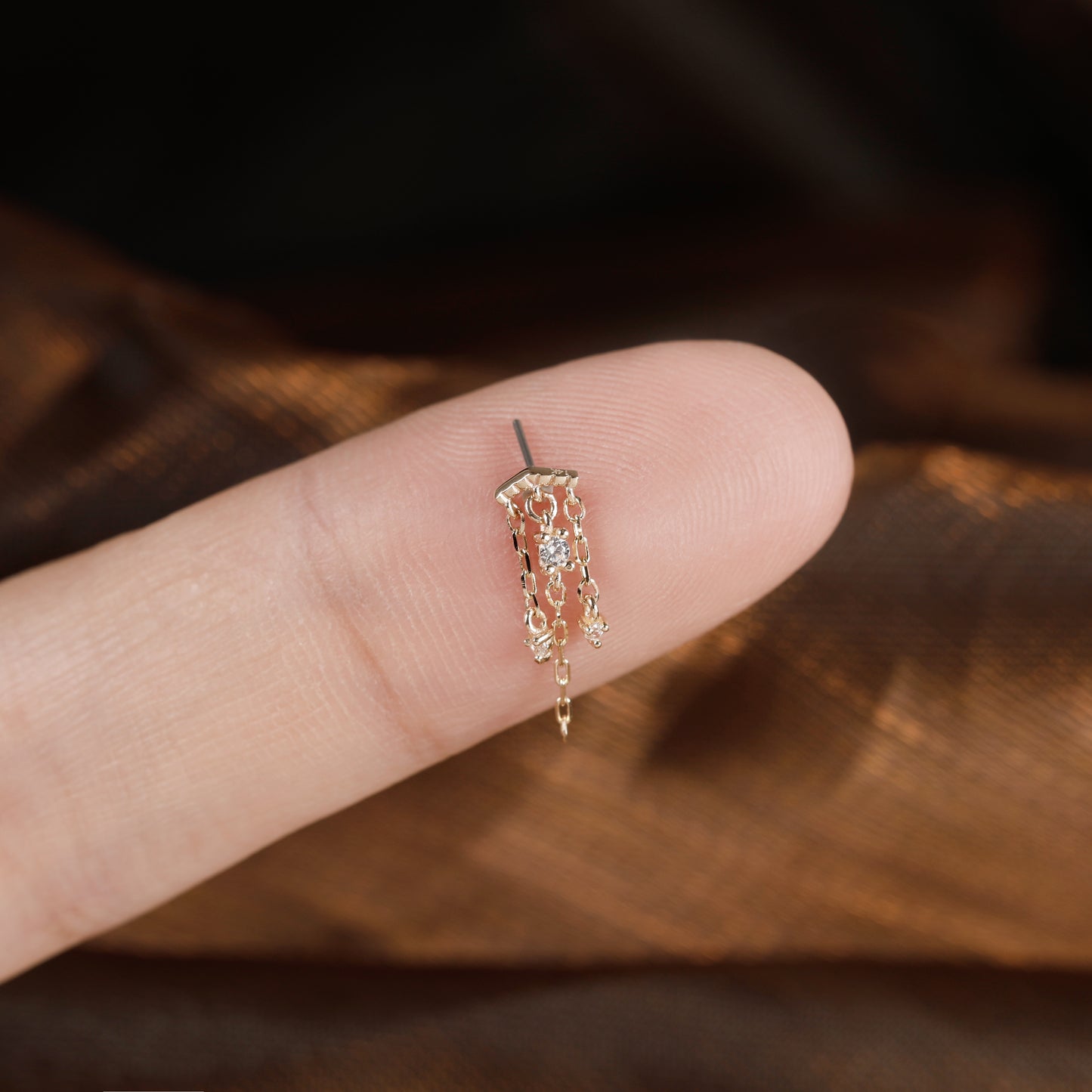 A delicate gold-toned dangle earring with small clear stones, shown resting on a person's fingertip for scale against a dark brown textured background.