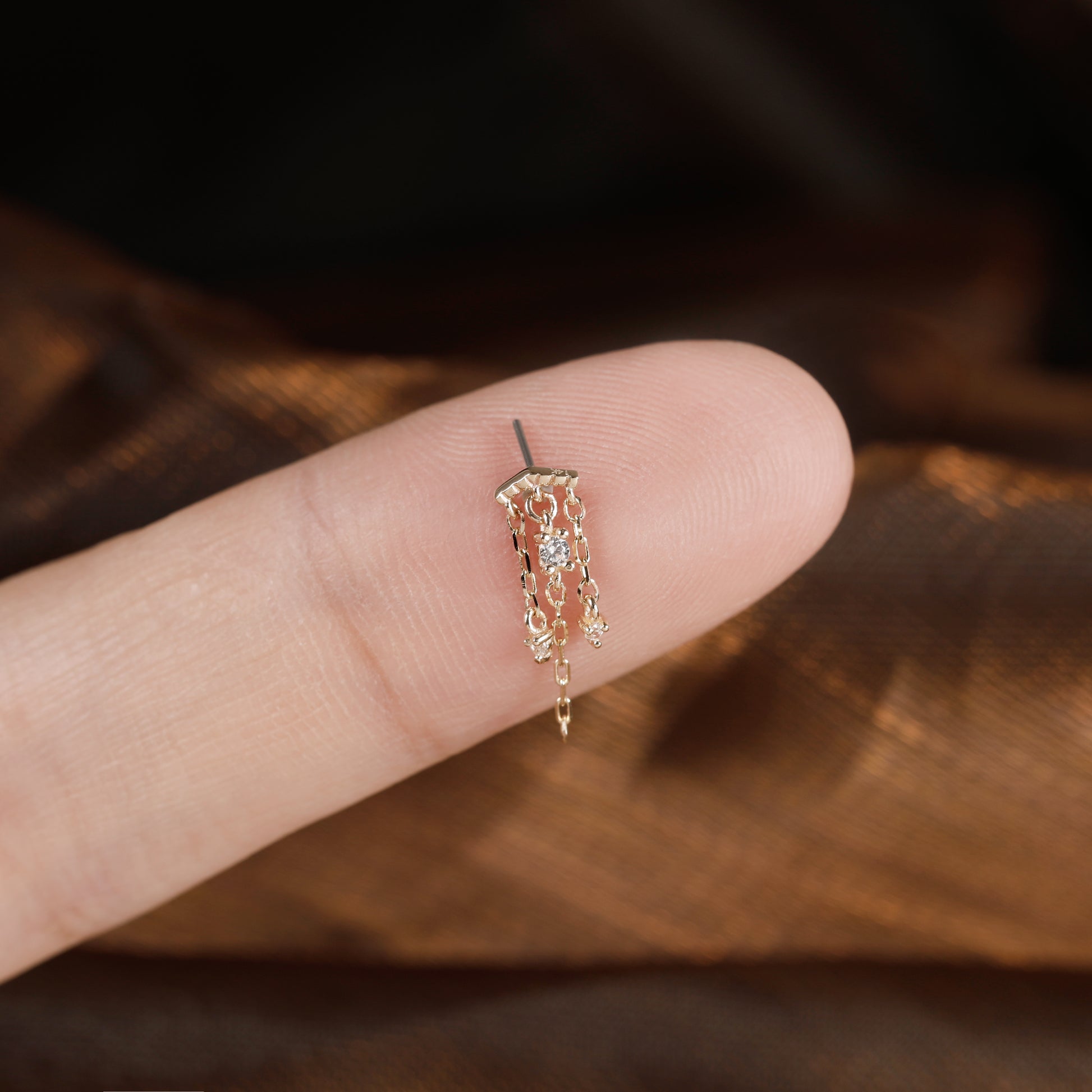A delicate gold-toned dangle earring with small clear stones, shown resting on a person's fingertip for scale against a dark brown textured background.