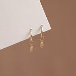 A pair of gold huggie hoop earrings with dangling hamsa hand charms, each accented with a small clear gemstone, displayed against a warm brown background and a white geometric card.