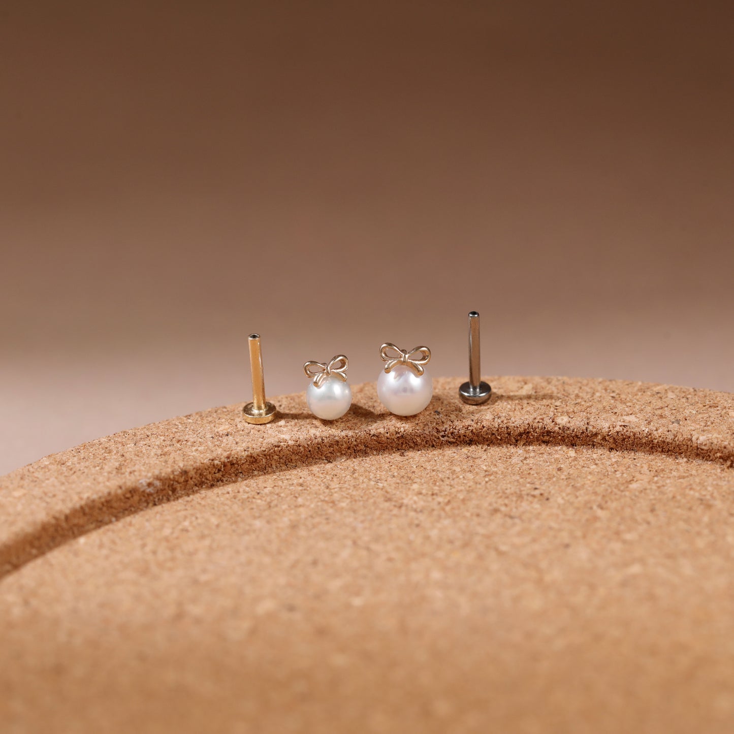 Three small pearl stud earrings with gold-toned posts displayed on a textured tan surface against a warm brown gradient background.