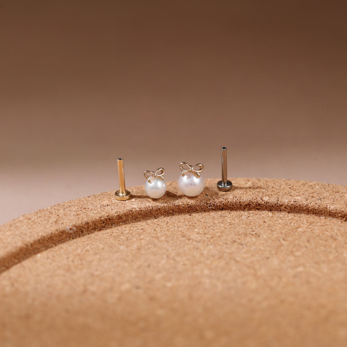 Three small pearl stud earrings with gold-toned posts displayed on a textured tan surface against a warm brown gradient background.
