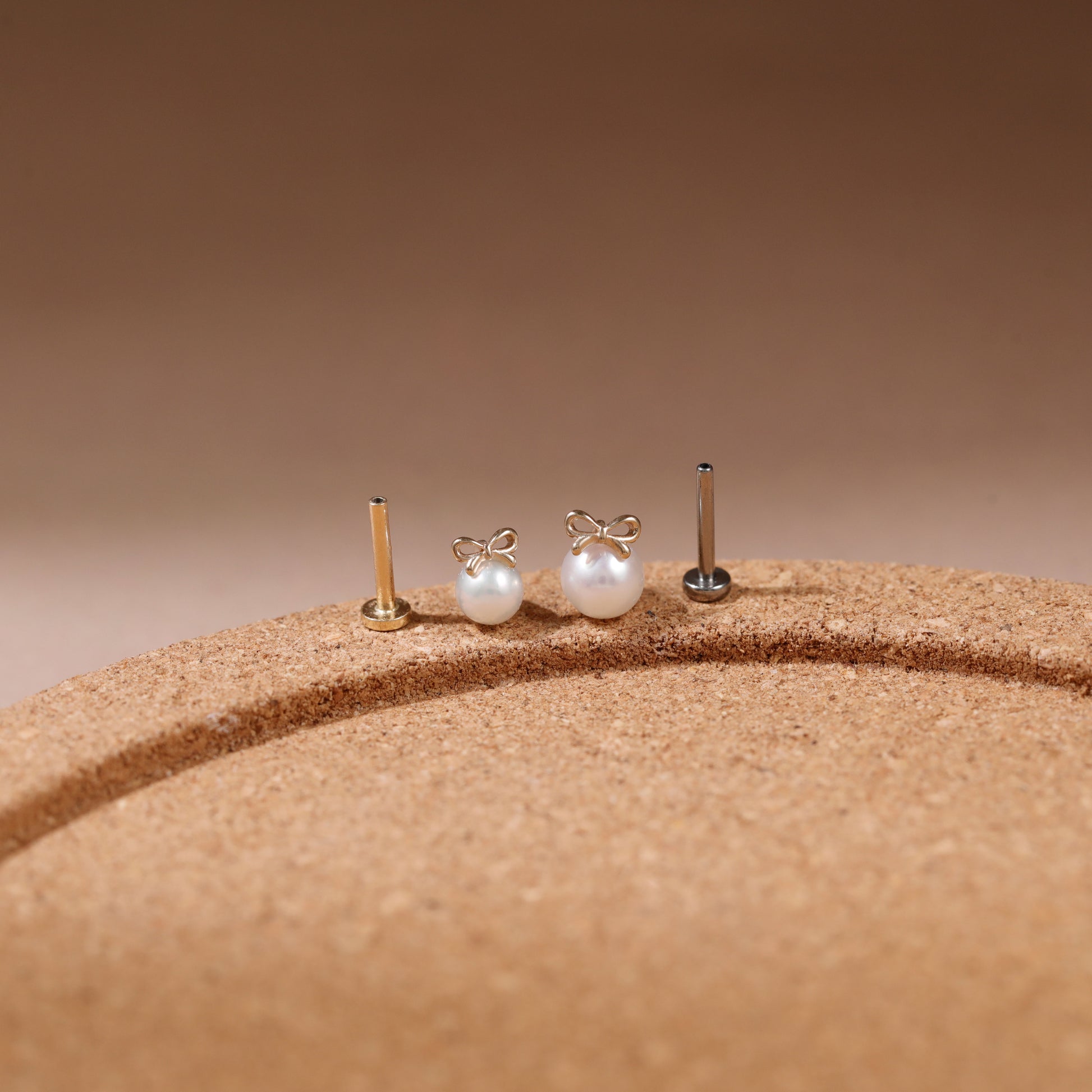 Three small pearl stud earrings with gold-toned posts displayed on a textured tan surface against a warm brown gradient background.