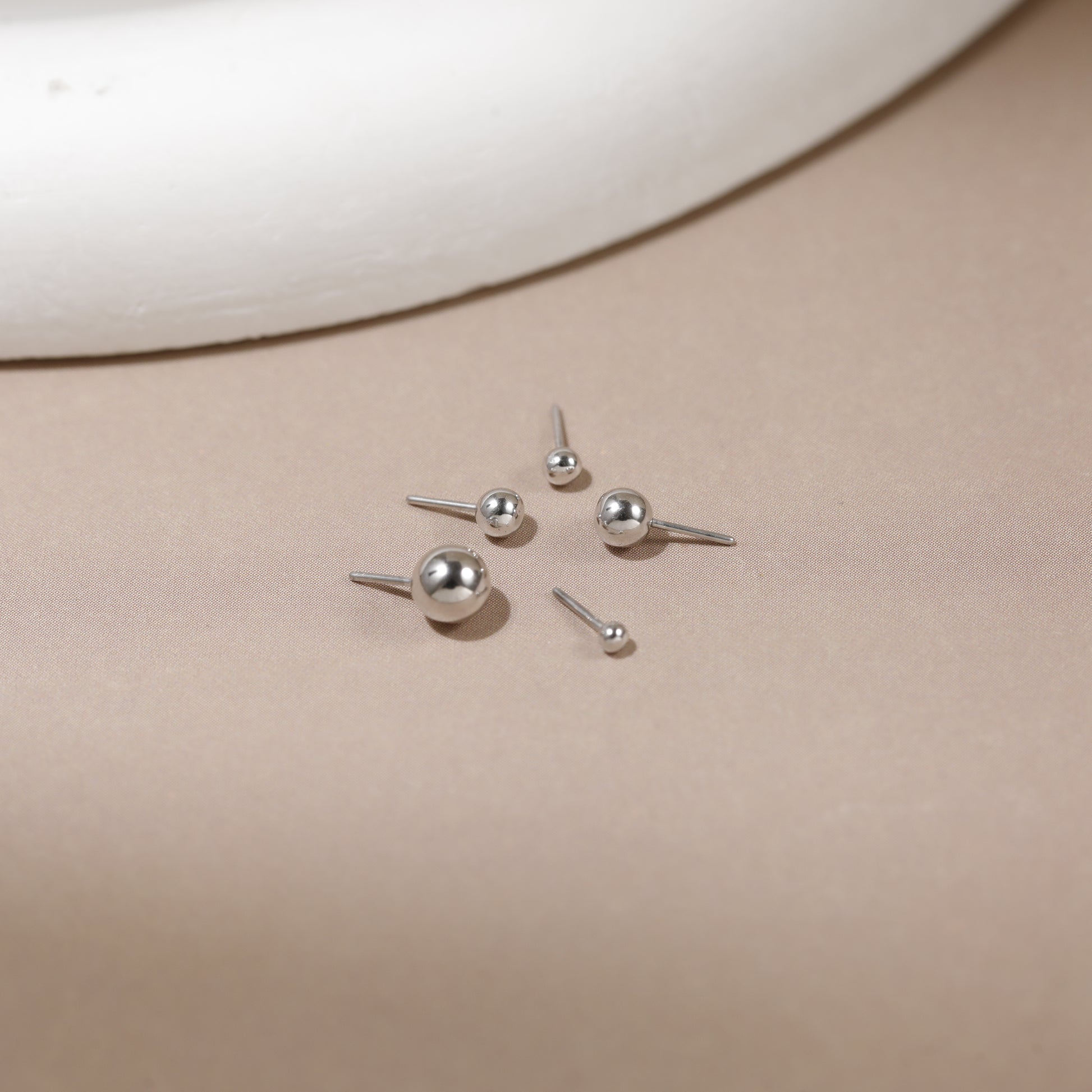 Five silver stud earrings with spherical ball tops in two sizes, arranged on a neutral beige surface with a white curved prop in the background.