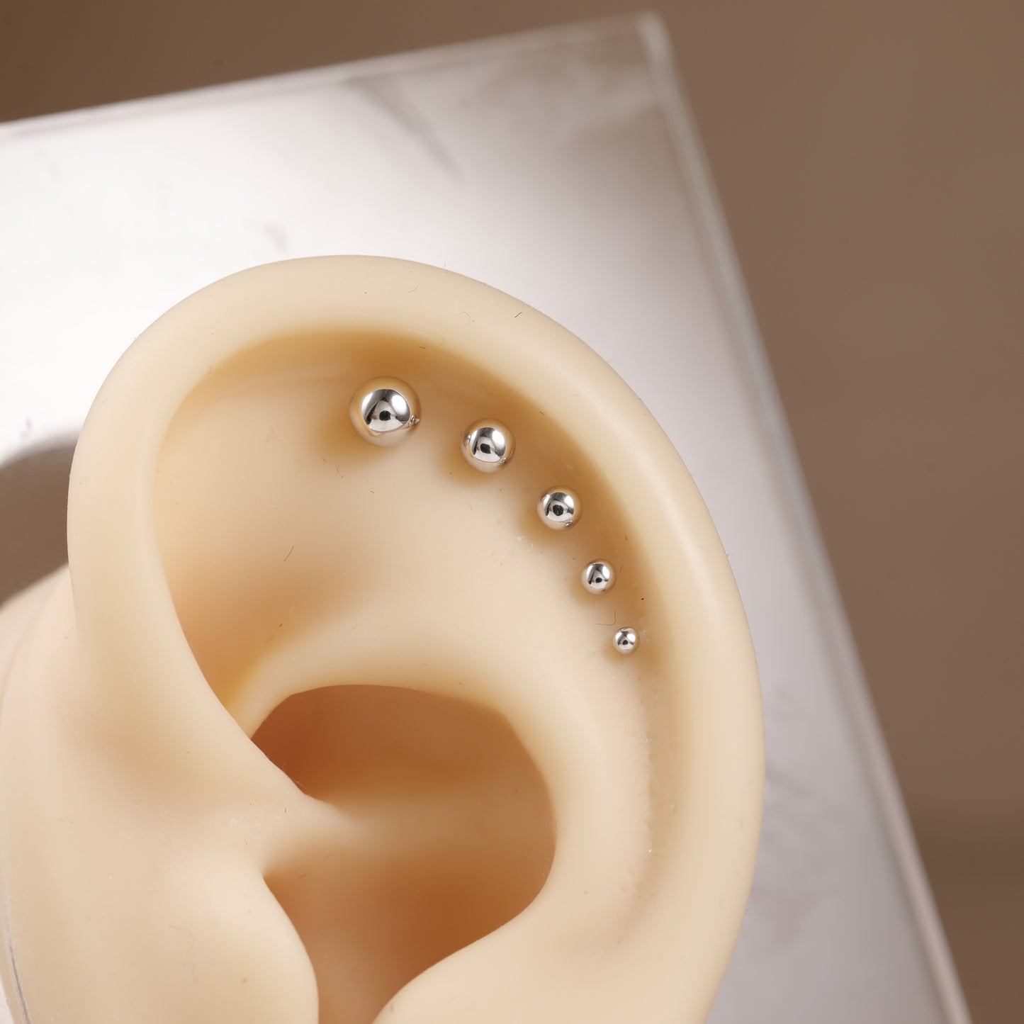 Five silver metallic ear barbells of varying sizes displayed on a beige silicone ear model, set against a neutral beige background.
