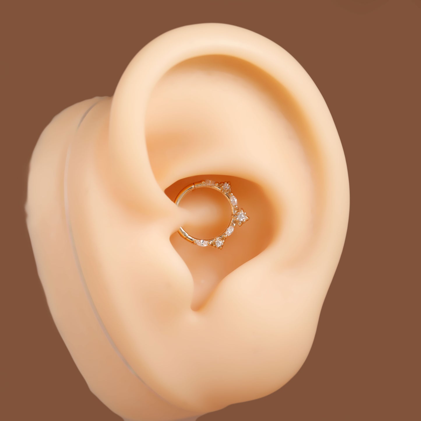 A silver-toned hoop ear piercing ring decorated with small clear crystals, shown on a beige silicone ear model against a solid brown background.