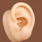 A silver-toned hoop ear piercing ring decorated with small clear crystals, shown on a beige silicone ear model against a solid brown background.