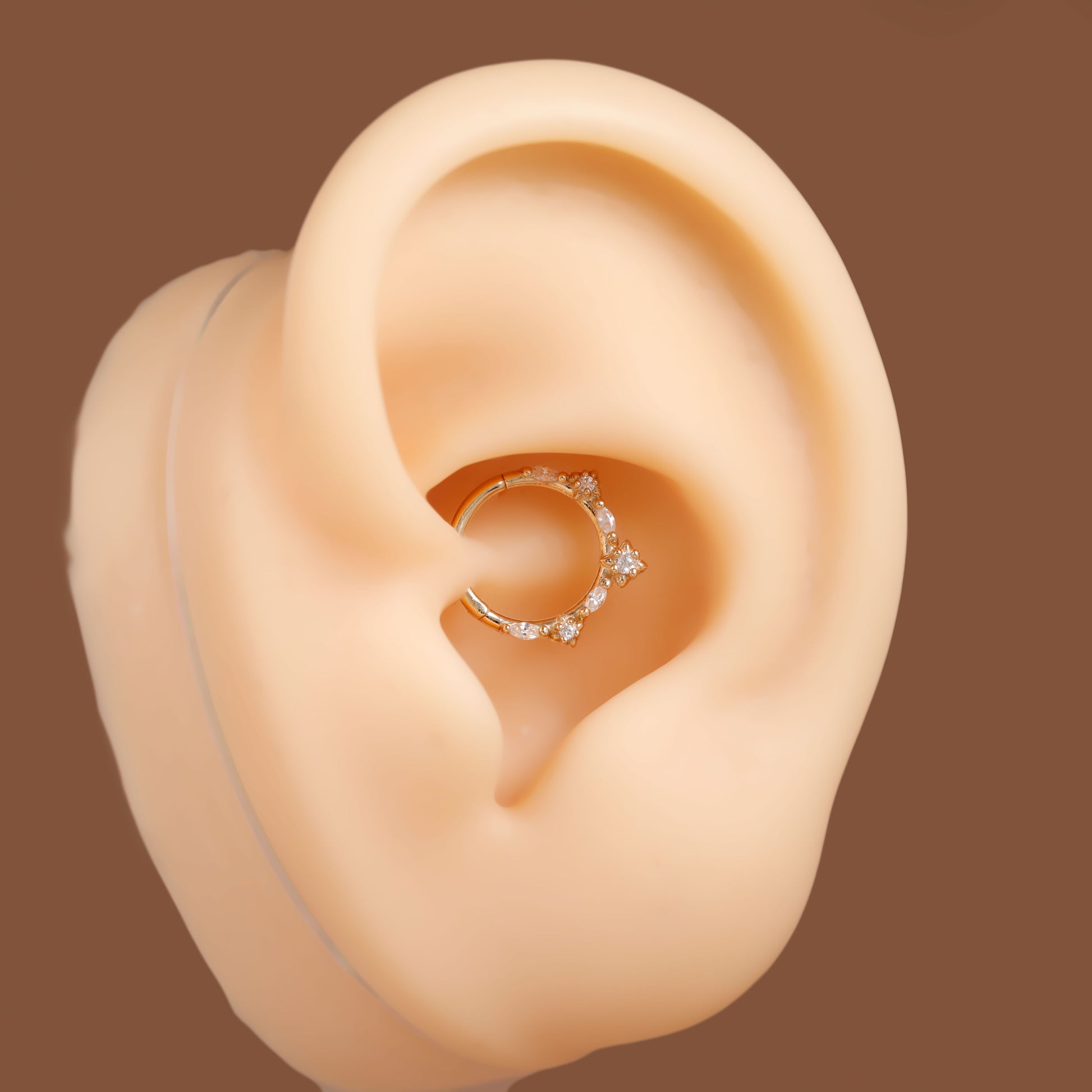 A silver-toned hoop ear piercing ring decorated with small clear crystals, shown on a beige silicone ear model against a solid brown background.