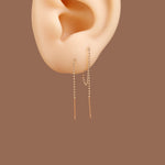 A pair of delicate gold dangle earrings with small bead chains and straight drop ends, shown worn on a beige silicone ear model against a solid brown background.