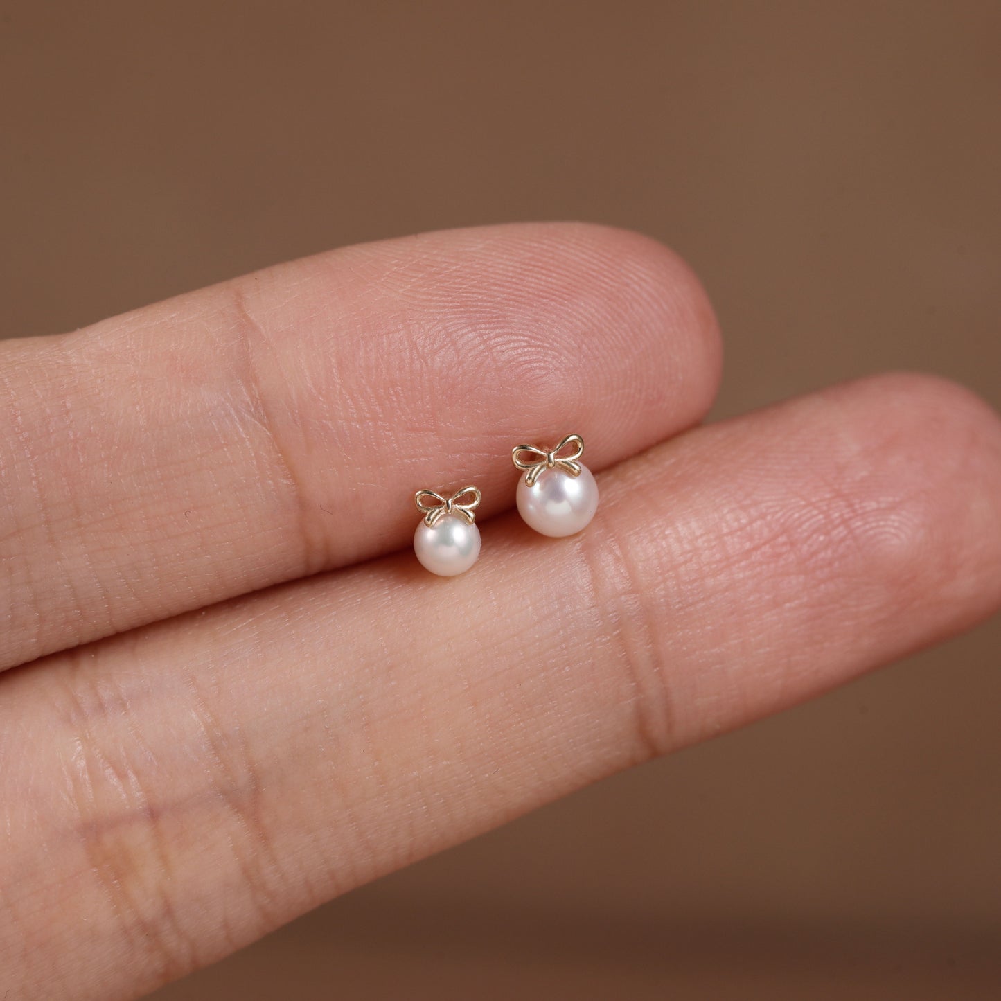 A pair of small pearl stud earrings with gold bow details, shown resting on a person's fingers against a soft brown background.