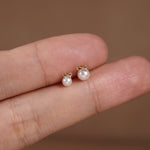 A pair of small pearl stud earrings with gold bow details, shown resting on a person's fingers against a soft brown background.
