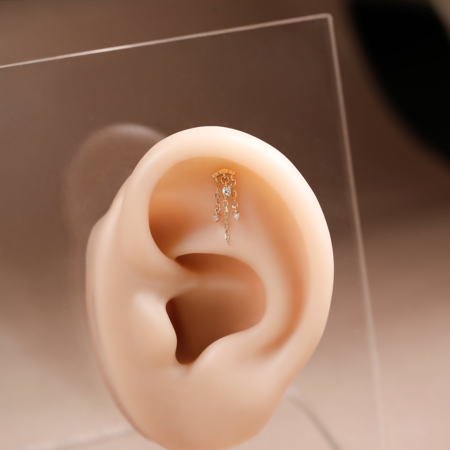 A small gold ear cuff with dangling clear stones displayed on a beige silicone ear model against a warm neutral background.