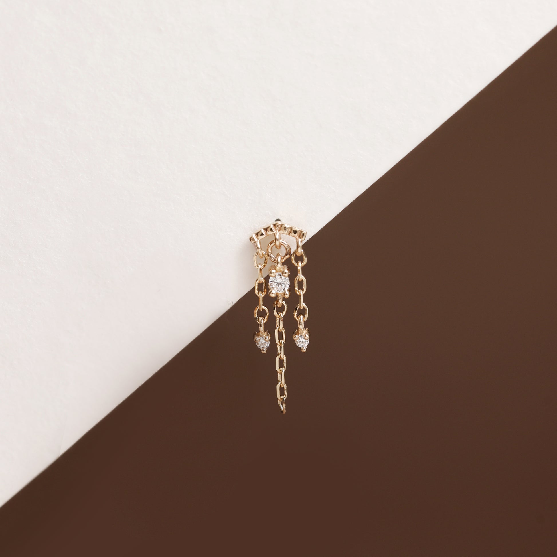 A delicate gold dangle earring with clear gemstones, resting on a diagonal split background of off-white and dark brown.