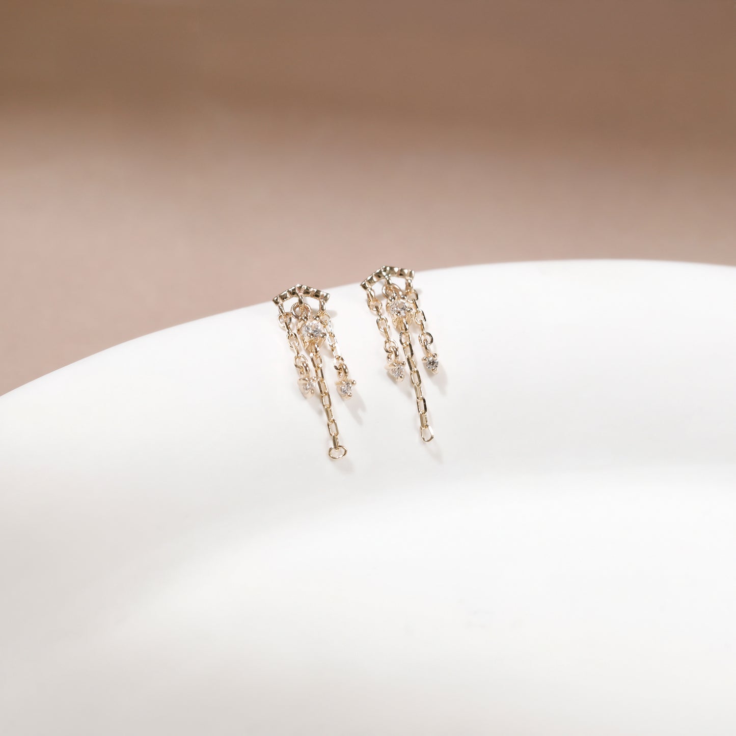A pair of delicate gold-toned dangle earrings with small clear stones and chain-like drops, displayed on a white curved surface against a soft beige background.
