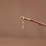 A delicate gold hamsa hand earring with a small clear gemstone, hanging from a thin brown twig against a soft, warm brown background.