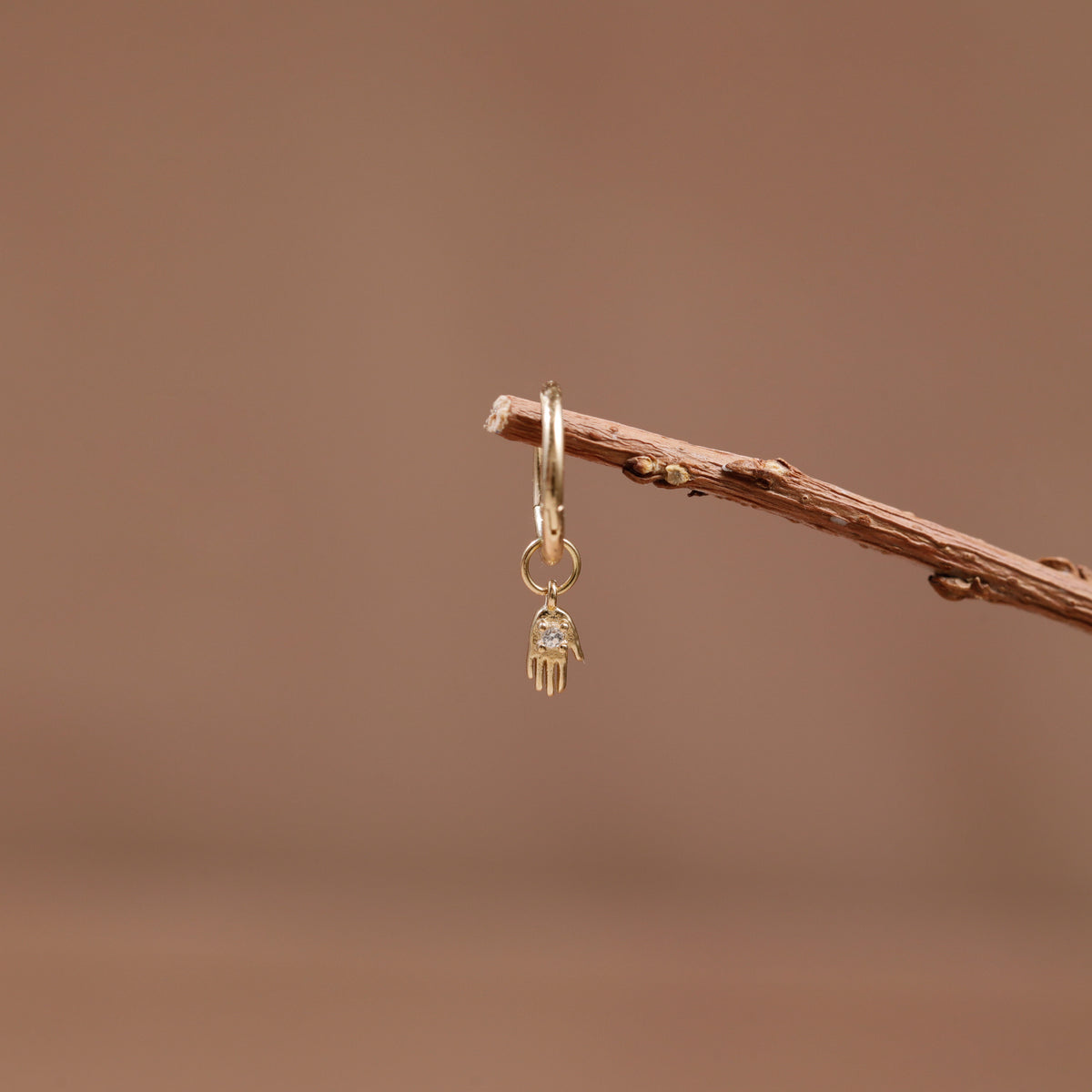 A delicate gold hamsa hand earring with a small clear gemstone, hanging from a thin brown twig against a soft, warm brown background.