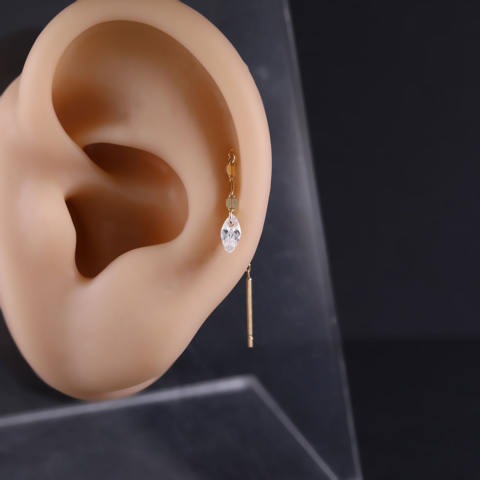 A gold hoop earring with a clear faceted drop and a thin dangling bar, shown on a beige ear mannequin against a dark background.
